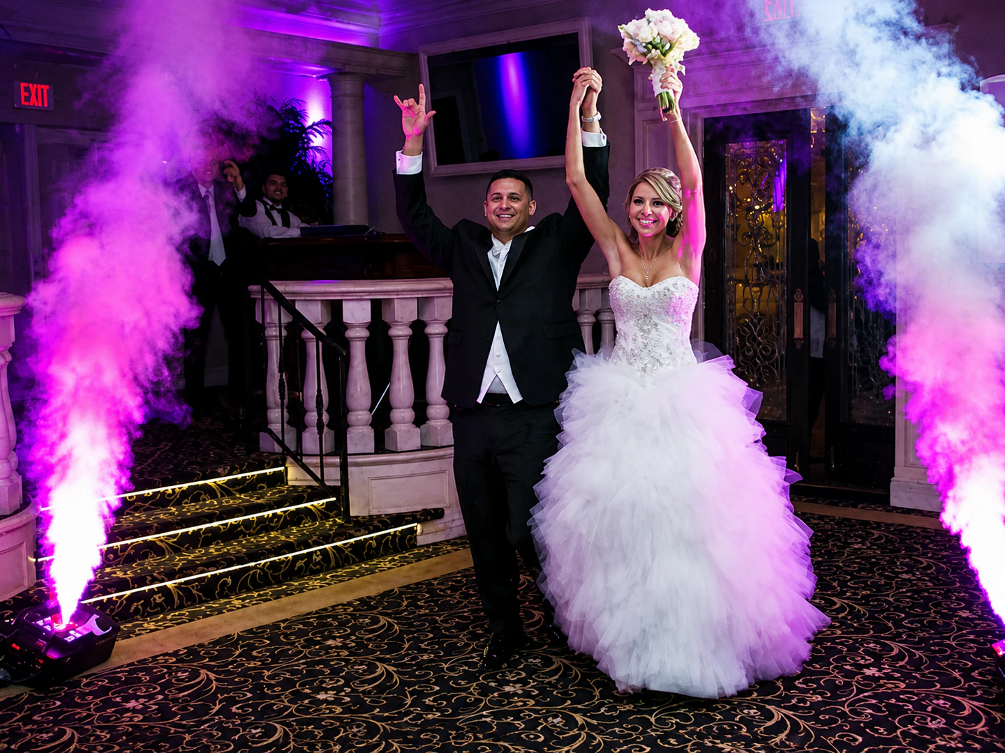 contentcreator - Stylus Weddings Bride in a strapless white gown and groom in a black tux raise their arms in celebration as purple smoke swirls around them inside a decorated venue.