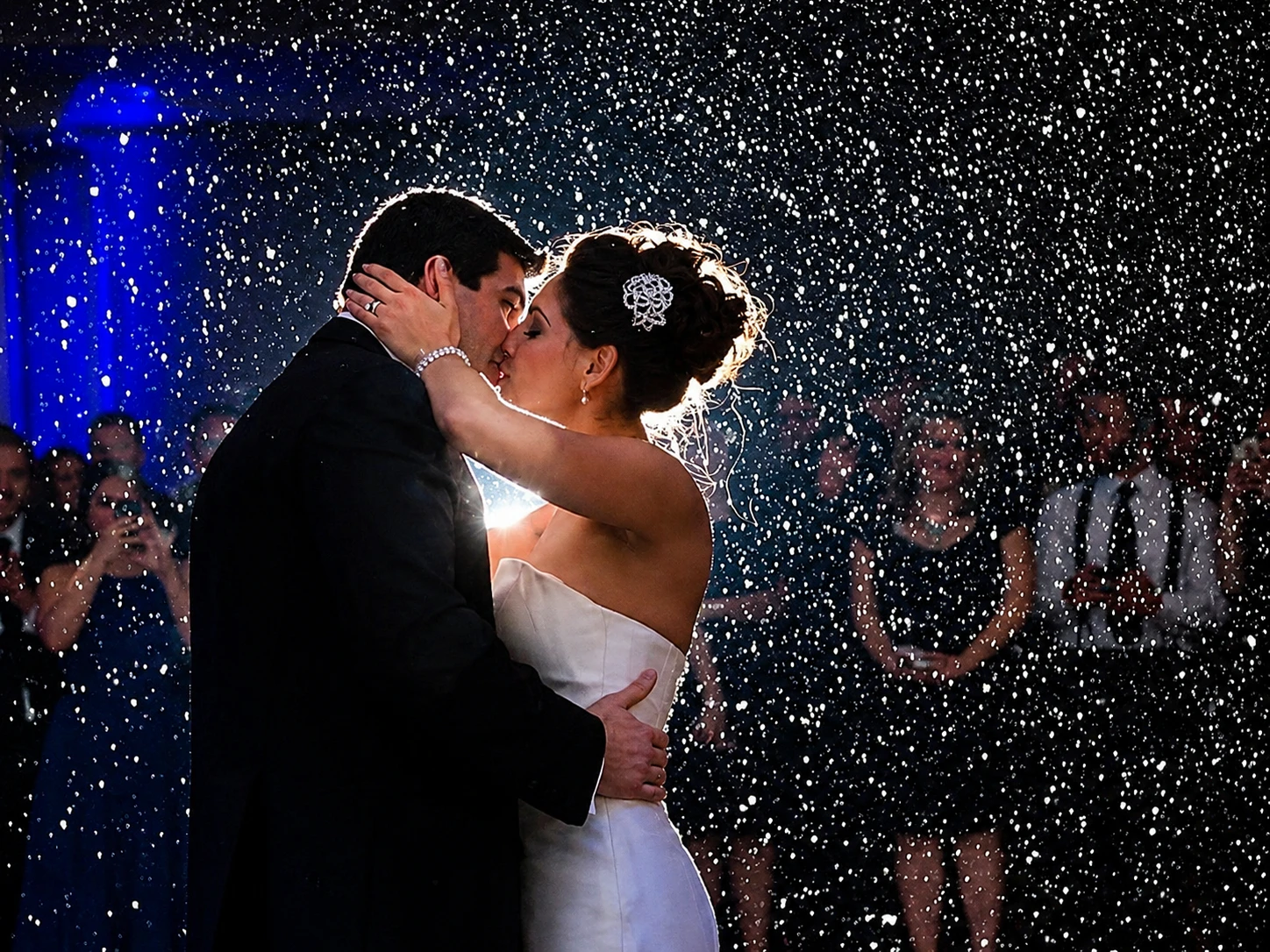 fakesnow - Stylus Weddings Bride and groom kiss while rain-like confetti falls around them at their wedding, guests blurred in the background, night scene.