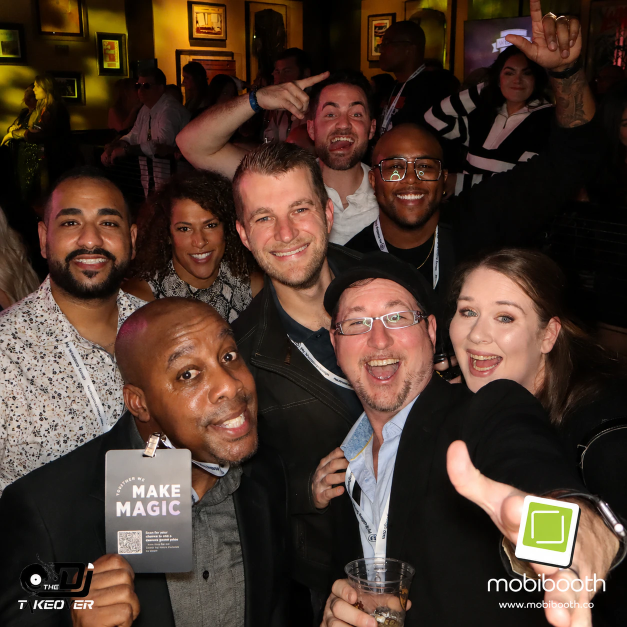 team - Stylus Weddings Group of smiling people posing for a selfie at a crowded party, with badges and a 'Make Magic' card visible in the foreground.
