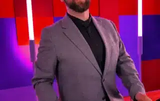 Smiling DJ in a gray blazer stands at a mixing console in a colorful studio with red and purple wall panels.