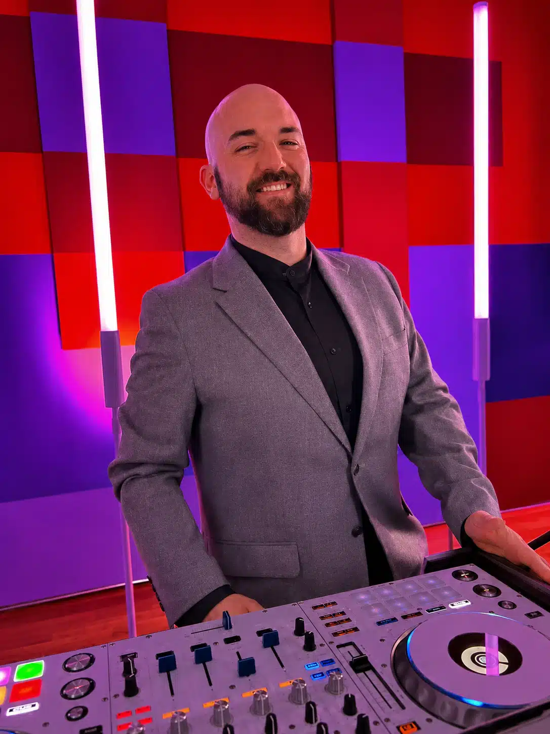 Smiling DJ in a gray blazer stands at a mixing console in a colorful studio with red and purple wall panels.